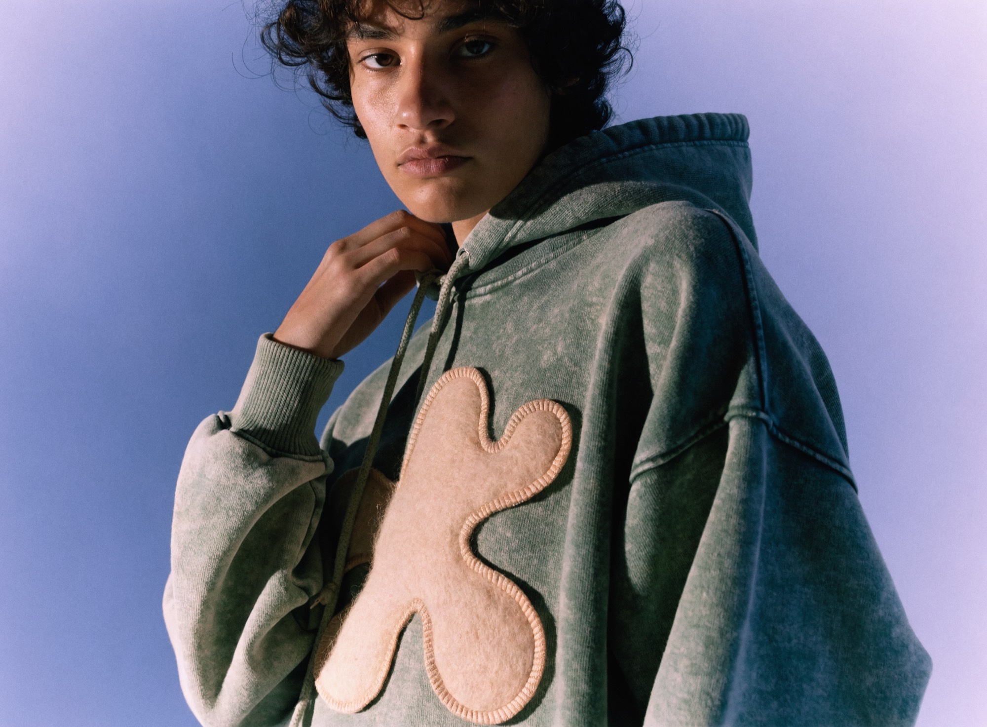 A sage pigment-washed hoodie with a raised appliqué patch, stitched at the edge